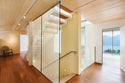 Modern interior staircase with glass railings and transparent glass panels, set in a minimalist home with light wood ceiling, dark wood flooring, and an outdoor lake view.