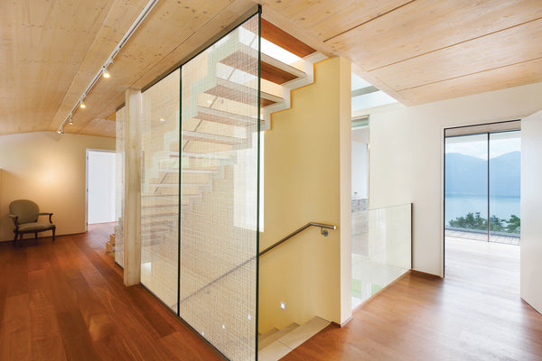 Modern interior staircase with glass railings and transparent glass panels, set in a minimalist home with light wood ceiling, dark wood flooring, and an outdoor lake view.